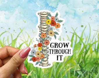 May include: A sticker featuring a stylized spine design adorned with colorful flowers in shades of orange, yellow, and blue. The text "GROW THROUGH IT" is displayed in a bold, black font. The sticker is held against a backdrop of grass and a blue sky.
