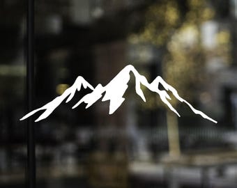 May include: A white mountain range silhouette. The design features three peaks of varying heights, with sharp, angular lines. The image is a simple, minimalist design, suitable for decals, stickers, or wall art. The background is blurred.