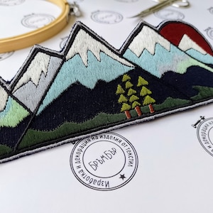 May include: Embroidered mountain range patch featuring snow-capped peaks, a setting sun, and evergreen trees. The patch has a dark blue border and is likely for clothing or accessories.