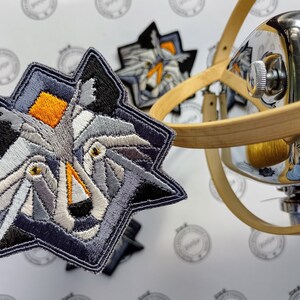 May include: A gray, white, and orange embroidered patch of a wolf's face. The patch is in the shape of a star and has a geometric design.