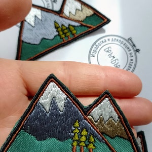 Puede incluir: Dos parches bordados para planchar que representan una cordillera con picos nevados, árboles verdes y un contorno marrón. Los parches están sobre un fondo blanco.