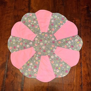 May include: A pink and gray floral patterned round table runner with a scalloped edge.