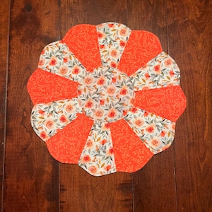 May include: A round, orange and white floral table runner with a scalloped edge. The runner is made of fabric and has a floral pattern with orange and white flowers.