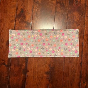 May include: A rectangular gray fabric placemat adorned with a repeating pattern of small, colorful flowers in pink, orange, yellow, and blue. The placemat is set against a dark wood surface.