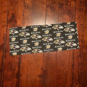 May include: A rectangular black fabric item featuring a repeating pattern of the Baltimore Ravens logo in purple, gold, and white. The logo includes a stylized raven and the team name. The item is displayed on a dark wood surface.
