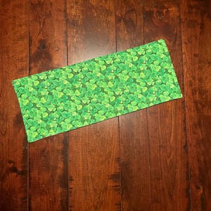 May include: A rectangular green fabric item with a repeating pattern of four-leaf clovers. The fabric is a bright, vibrant green, and the clovers are in varying shades of green. The item is placed on a dark brown wooden surface.