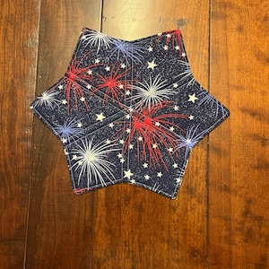 May include: A star-shaped fabric coaster with a navy blue background. The coaster features a pattern of red, white, and blue fireworks and white stars. The coaster is likely used for placing drinks on a table to protect the surface.