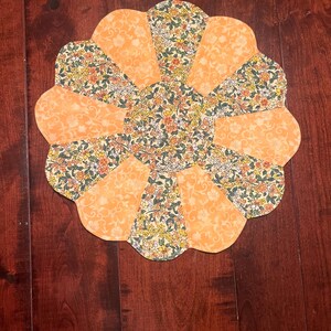 Puede incluir: Un salvamanteles acolchado hecho a mano con forma de flor. El salvamanteles presenta secciones alternas de tela naranja lisa y tela estampada floral con flores amarillas, verdes y naranjas. El salvamanteles está colocado sobre una superficie de madera oscura.