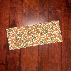 May include: Rectangular placemat featuring a cream backdrop adorned with a repeating design of orange pinecones and green foliage. This fabric placemat is intended for table settings. The design evokes an autumnal or fall theme.