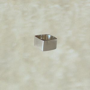 May include: A silver-coloured, square-shaped ring. The ring has a solid, geometric design with a flat top and sides. The ring is made of a shiny, reflective metal, and the interior is hollow. The ring is a piece of jewellery.