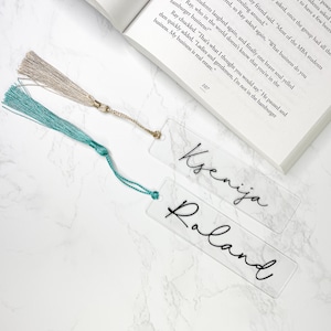 May include: Two clear acrylic bookmarks with black calligraphy. The bookmarks have tassels attached. The top bookmark reads "Ksenija" and the bottom bookmark reads "Roland".