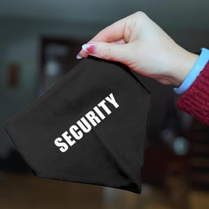 May include: A black triangular bandana with the word "SECURITY" printed in large white capital letters. The bandana is held by a hand with red nail polish, and the background is blurred.