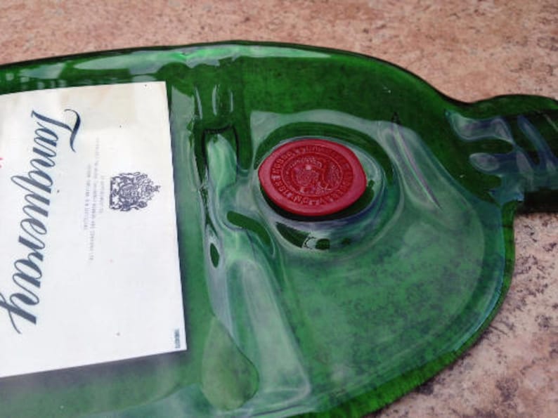 Op de afbeelding: Een groene glazen fles met een etiket waarop "Gordon's" staat en een rode waszegel. De fles is leeg en heeft een getextureerd oppervlak.