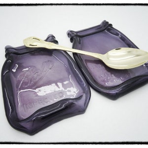 May include: Two purple glass spoon rests shaped like Ball mason jars with the words "Ball" and "Improved" embossed on the front. A gold spoon is resting on one of the spoon rests.