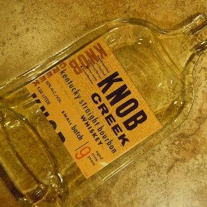 May include: A clear glass bottle with a textured surface. The bottle is labeled "Knob Creek Kentucky Straight Bourbon Whiskey" with the details "50% Alc/Vol 1.00 Liter Small Batch Aged Nine Years".