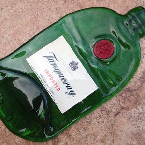Pode incluir: Uma garrafa de vidro verde com um r&oacute;tulo que diz "Tanqueray Imported" e um selo de cera vermelho. A garrafa tem a forma de uma garrafa de gin tradicional e tem uma superf&iacute;cie texturizada.