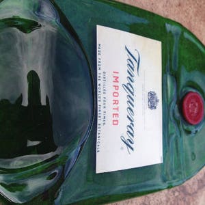 Op de afbeelding: Een groene glazen fles met een etiket waarop staat "Tanqueray Ge&iuml;mporteerd Viermaal Gedistilleerd Gemaakt van 's werelds beste botanicals". De fles heeft een rode dop en een doorzichtige glazen stop.