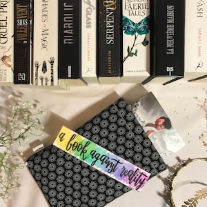 Peut inclure: Une collection de livres avec un marque-page indiquant "a book against reality". Le marque-page est coloré avec un motif aquarelle. Les livres sont sur une surface blanche avec une couverture de livre à motifs noir et blanc.