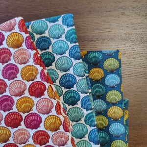 Peut inclure: Trois morceaux de tissu avec des motifs de coquillages. L'un a un fond blanc avec des coquillages roses, oranges et jaunes. Un autre a un fond blanc avec des coquillages bleus et turquoise. Le troisième a un fond bleu foncé avec des coquillages verts et jaunes.