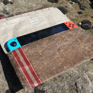 Peut inclure: Une housse en toile marron avec des rayures rouges et blanches, conçue pour contenir une console Nintendo Switch. La housse porte une étiquette indiquant "FLAXELIA HANDMADE GOODS".