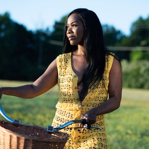 May include: A woman in a yellow crochet dress is holding a bicycle with a wicker basket. She is standing in a field of yellow flowers.