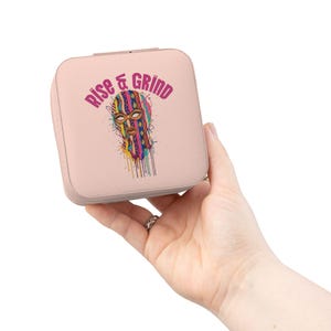 May include: A square, light pink jewelry travel case featuring the text "RISE & GRIND" in magenta above a colorful, stylized illustration of a person wearing a patterned balaclava with vibrant paint drip effects in shades of blue, yellow, and red.