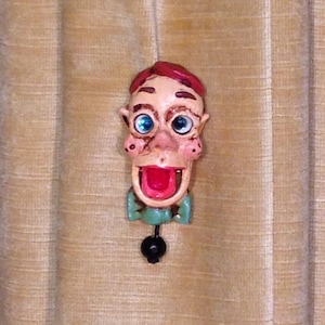 May include: A whimsical ceramic figurine of a man with a red hat, blue eyes, and a green bow tie. The figurine has a large, open mouth and a playful expression.