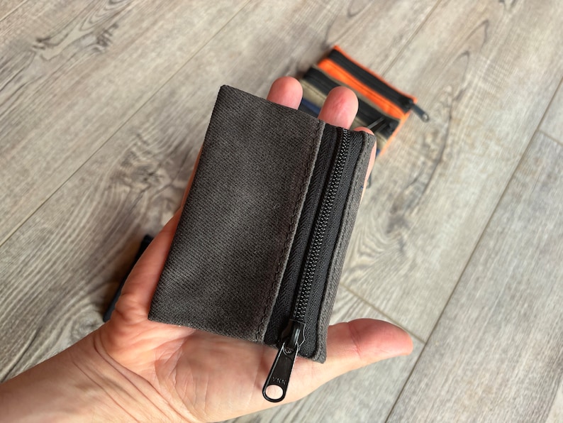 May include: A small gray zippered pouch with a black zipper. The pouch is made of a soft, textured fabric.