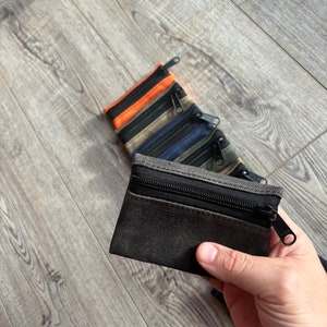 May include: A black canvas zippered pouch with a black zipper pull. The pouch is held in a hand. There are other pouches in the background, in orange, beige, blue, and green.