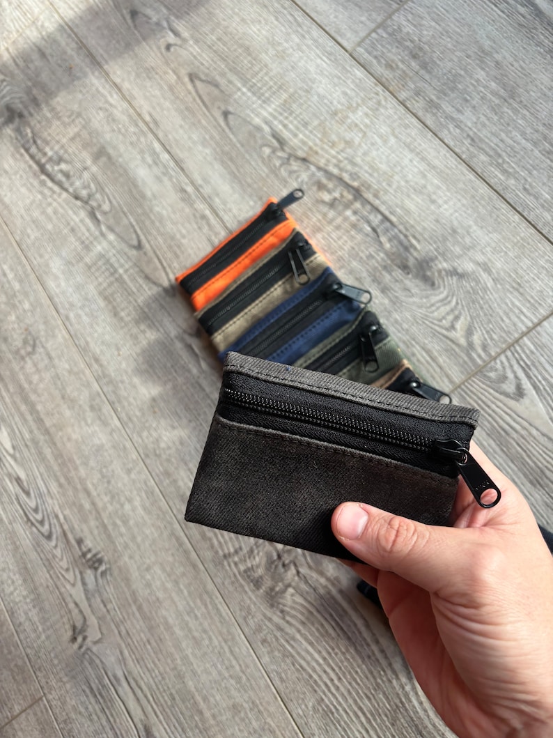 May include: A black canvas zippered pouch with a black zipper pull. The pouch is held in a hand. There are other pouches in the background, in orange, beige, blue, and green.