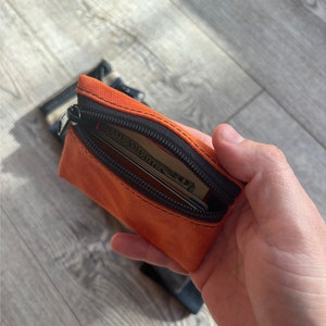 May include: A small orange zippered pouch with a black zipper and black trim. The pouch is open and has a credit card and a dollar bill inside.