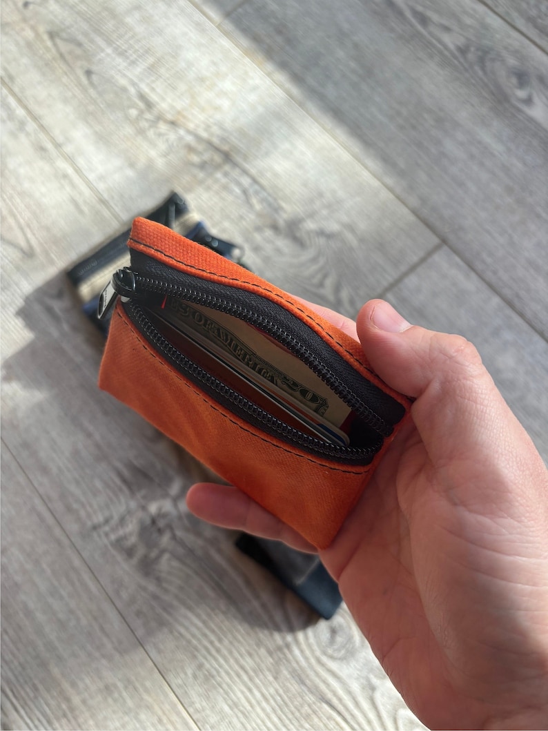 May include: A small orange zippered pouch with a black zipper and black trim. The pouch is open and has a credit card and a dollar bill inside.