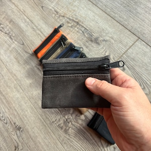 May include: A set of four small zippered pouches in various colors, including black, gray, orange, and blue. The pouches are made of a soft, textured fabric and have a zipper closure.