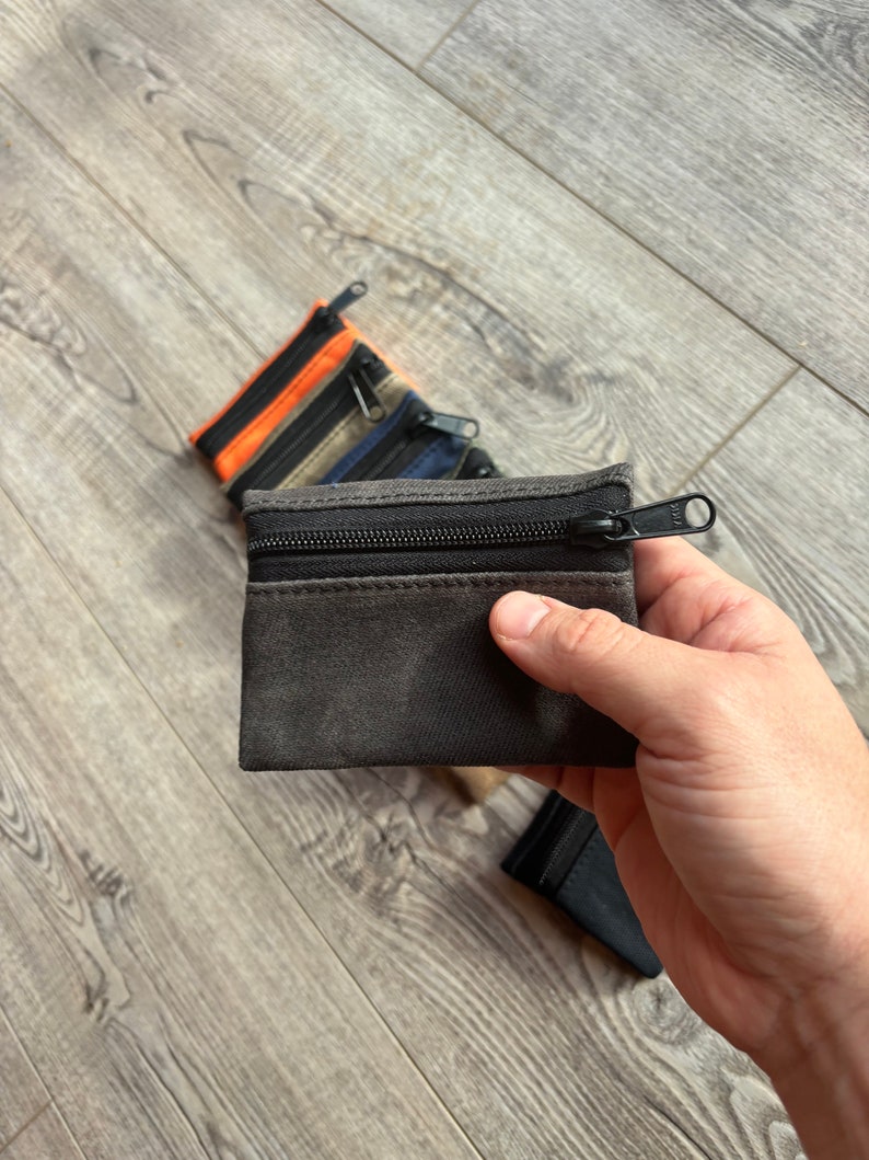 May include: A set of four small zippered pouches in various colors, including black, gray, orange, and blue. The pouches are made of a soft, textured fabric and have a zipper closure.