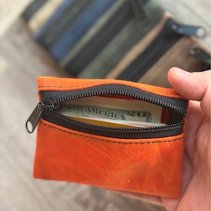 May include: A small orange zippered pouch with a black zipper and a dollar bill inside. The pouch is made of a soft, textured fabric.