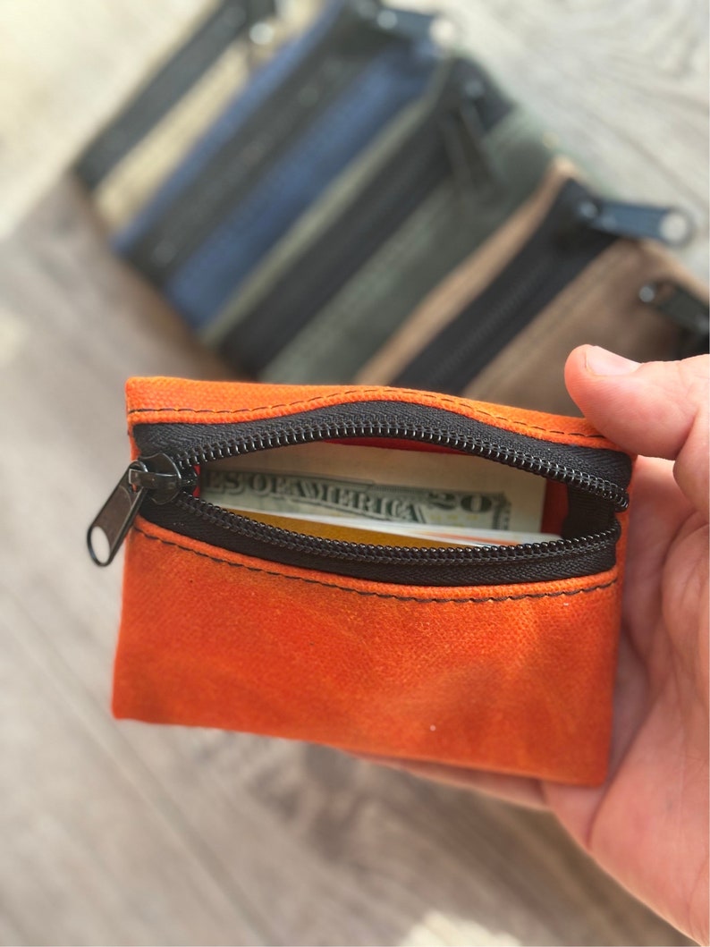 May include: A small orange zippered pouch with a black zipper and a dollar bill inside. The pouch is made of a soft, textured fabric.