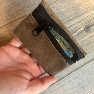 May include: A small brown canvas zipper pouch with a black zipper and a credit card and cash inside.