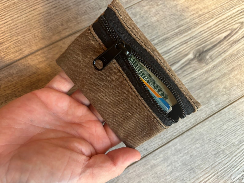 May include: A small brown canvas zipper pouch with a black zipper and a credit card and cash inside.