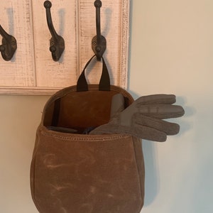 May include: A brown canvas bag with a black strap hanging on a white wooden hook rack. The bag is filled with a pair of gray gloves.