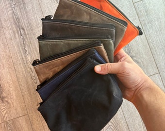 May include: A stack of six zippered pouches in various shades of brown, green, blue, and orange. The pouches are made of a canvas-like material and have a zipper closure.