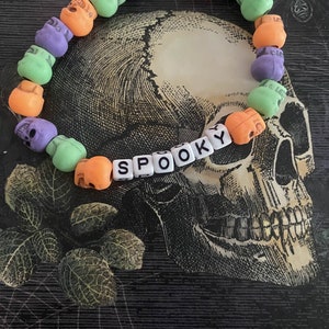 Op de afbeelding: Een kleurrijke armband gemaakt van plastic schedel kralen in tinten groen, oranje en paars. De armband spelt "SPOOKY" in witte letters.