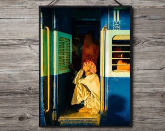Puede incluir: Un hombre está sentado en la puerta de un vagón de tren, mirando hacia abajo. El vagón de tren está pintado de azul y verde con detalles amarillos. El hombre lleva un turbante blanco y una camisa blanca.