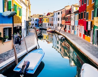 Può includere: Un canale stretto a Venezia, Italia, fiancheggiato da edifici colorati. L'acqua riflette gli edifici e il cielo. Due piccole barche sono ormeggiate nel canale.