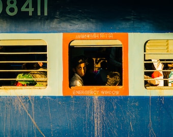 May include: A blue train car with three windows. The middle window is labeled "Emergency Window" in orange letters. A young person with dark hair is looking out the window. The windows have metal bars.