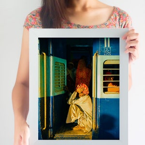 May include: A man sits on the floor of a train car, wearing a white turban and a white robe. The train car is painted blue and has yellow accents. The man is looking down at his hands.
