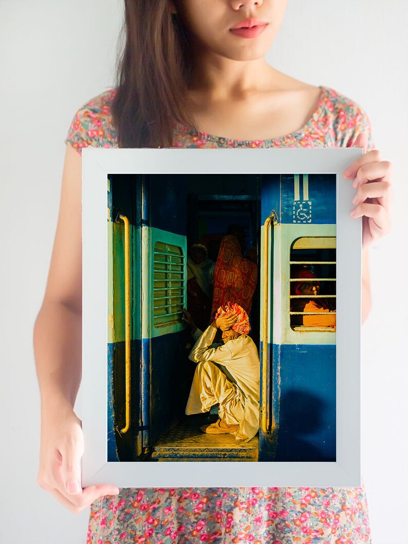May include: A man sits on the floor of a train car, wearing a white turban and a white robe. The train car is painted blue and has yellow accents. The man is looking down at his hands.