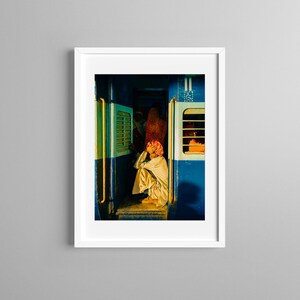 May include: A man sits on the floor of a train car, looking down. The train car is painted blue and has a window with a yellow frame. The man is wearing a white shirt and a red turban.