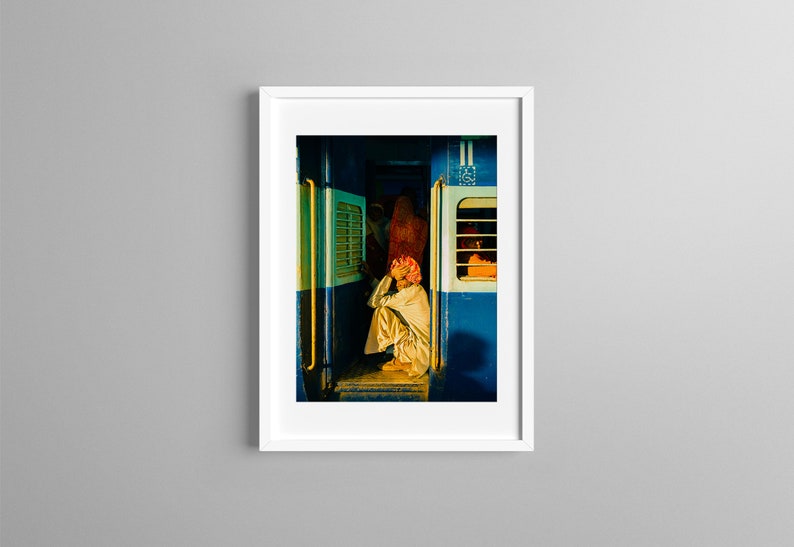 May include: A man sits on the floor of a train car, looking down. The train car is painted blue and has a window with a yellow frame. The man is wearing a white shirt and a red turban.