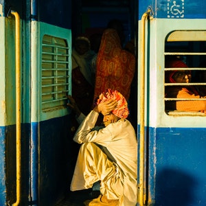 May include: A man wearing a yellow turban sits on the steps of a blue train car. The train car has a window with a barred grate and a sign with the Indian flag.