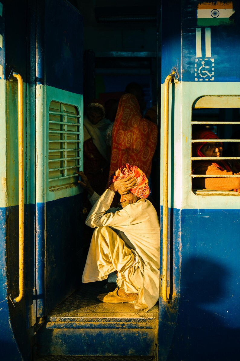 May include: A man wearing a yellow turban sits on the steps of a blue train car. The train car has a window with a barred grate and a sign with the Indian flag.